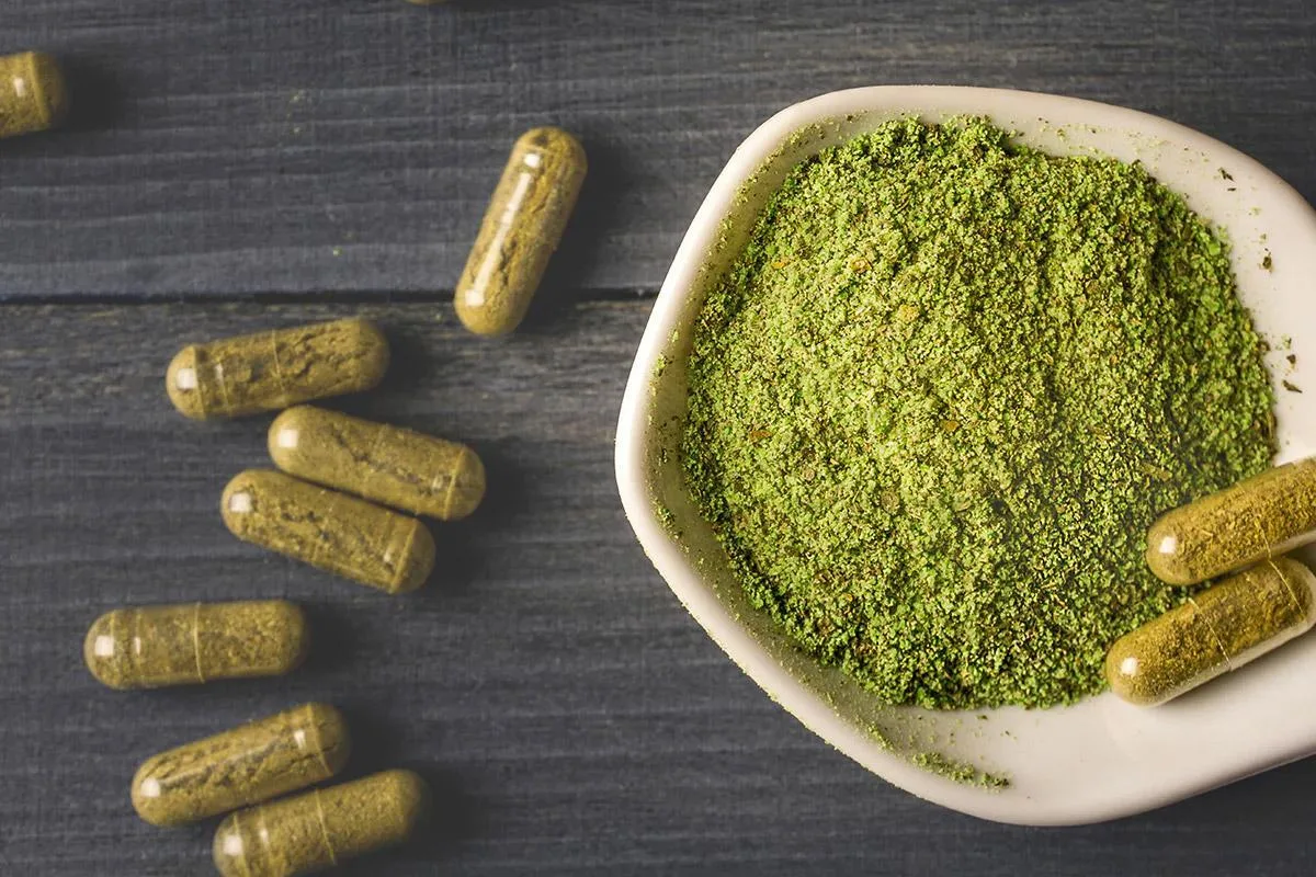 Green powder in a bowl with capsules on dark surface.